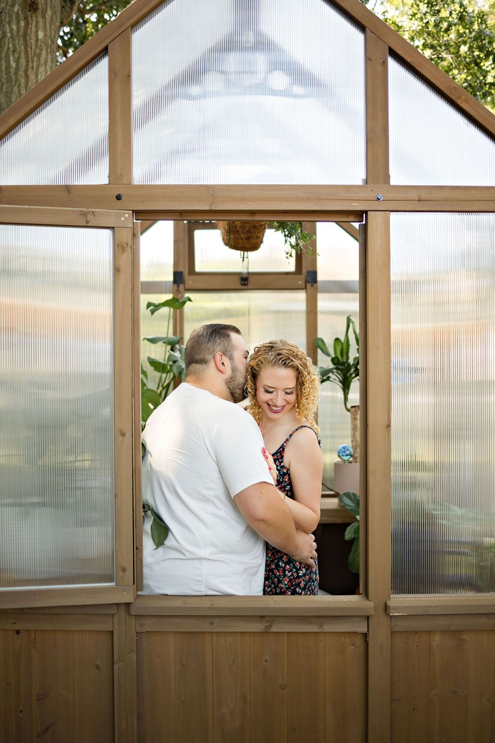 Couple embracing and smiling inside a wooden greenhouse with plants and polycarbonate panels on a sunny day.