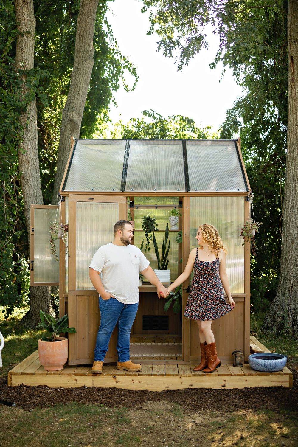 Couple holding hands in front of a wooden greenhouse surrounded by trees in a lush backyard garden.