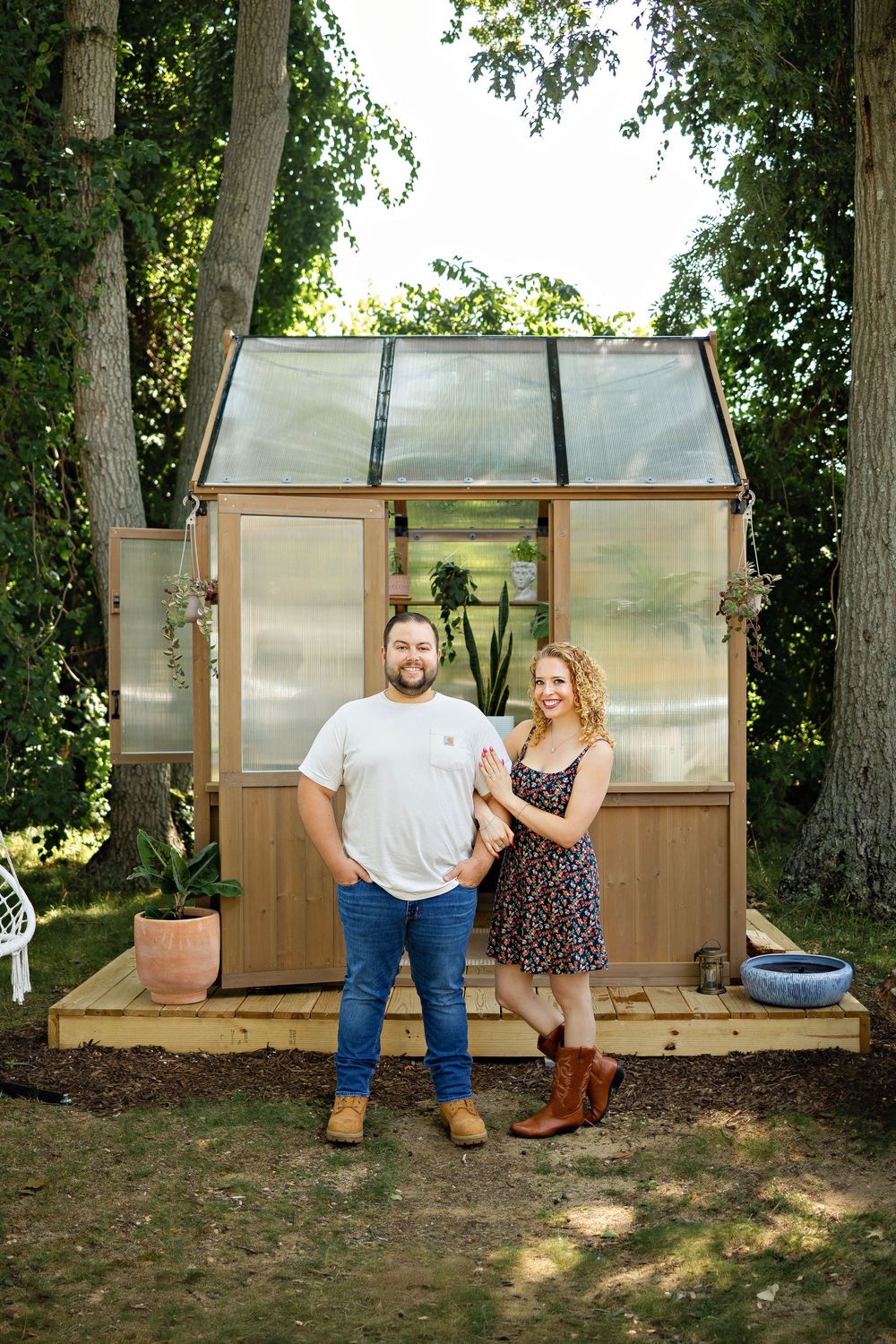 A couple stands smiling in front of a wooden greenhouse surrounded by trees in a lush backyard garden setting.