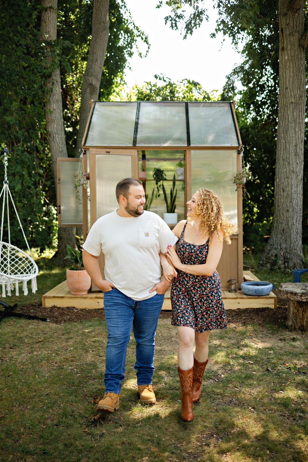 A couple holds hands smiling in a backyard garden with a glass greenhouse and lush trees behind them.