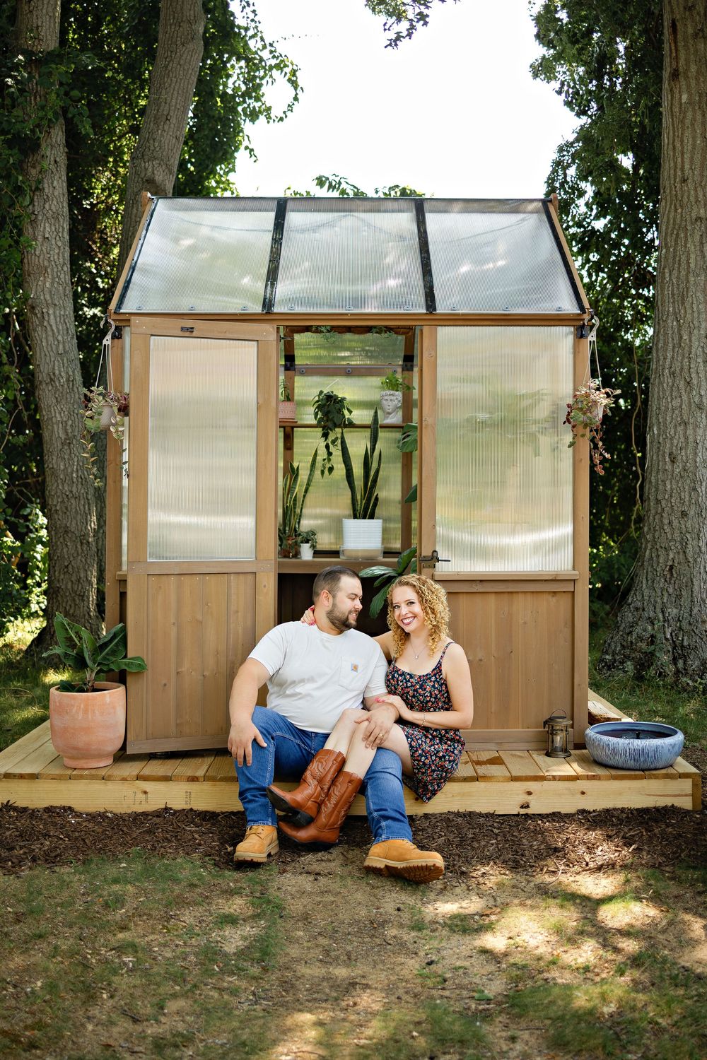A couple sits together smiling in front of a small wooden greenhouse surrounded by tall trees in a garden setting.