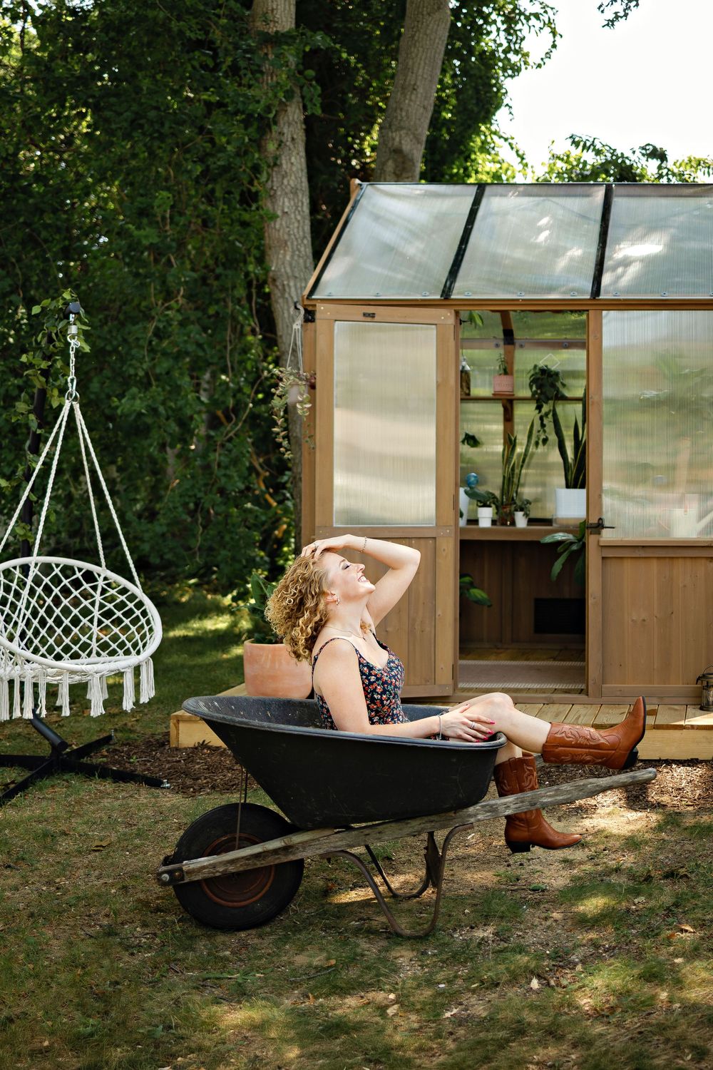 Woman relaxing in a wheelbarrow beside a wooden greenhouse with a macrame swing chair in a lush garden.