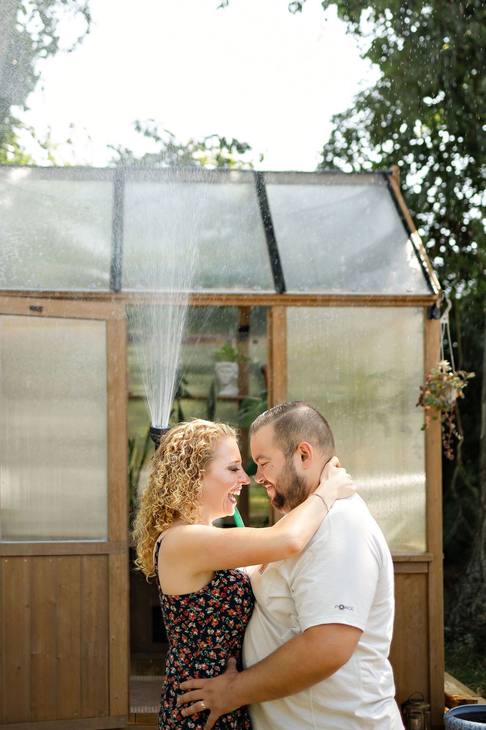 A couple embraces and gazes at each other tenderly in front of a rustic greenhouse with water misting in the background.