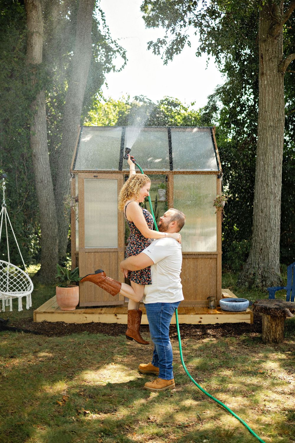 Couple dancing playfully in backyard near wooden greenhouse, woman holding hose, surrounded by lush trees on sunny day.
