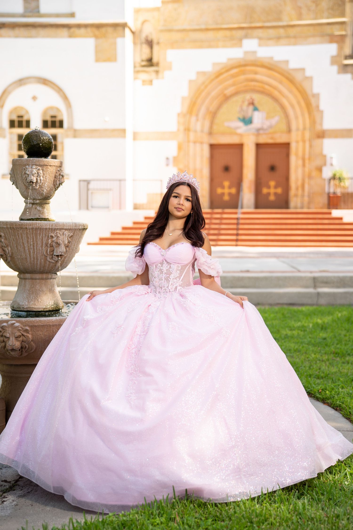 Young woman in pink quinceañera ball gown and tiara posing in front of a church with fountain.