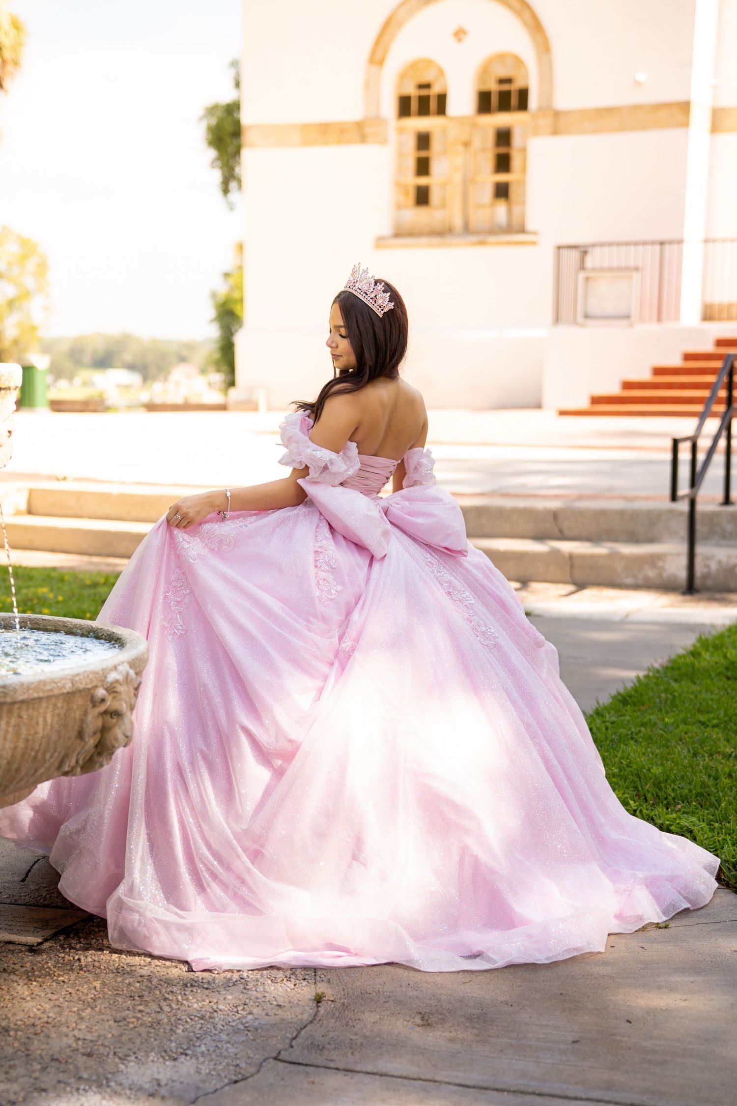 Young woman in a pink ball gown and tiara poses gracefully by a fountain outside an elegant building.