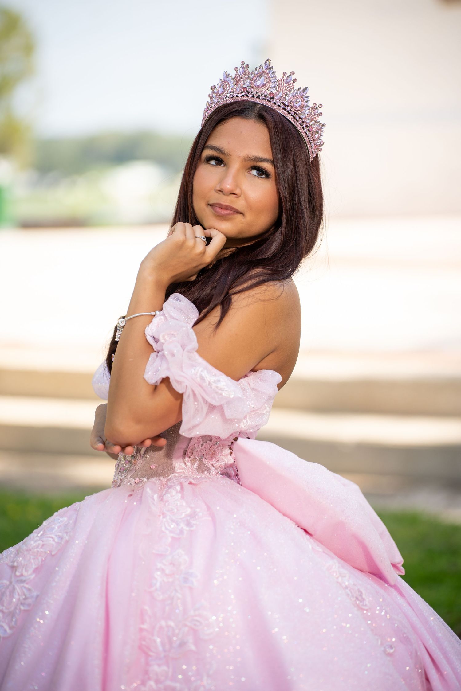Young Latina girl in pink ball gown and tiara poses gracefully for quinceañera portrait outdoors.