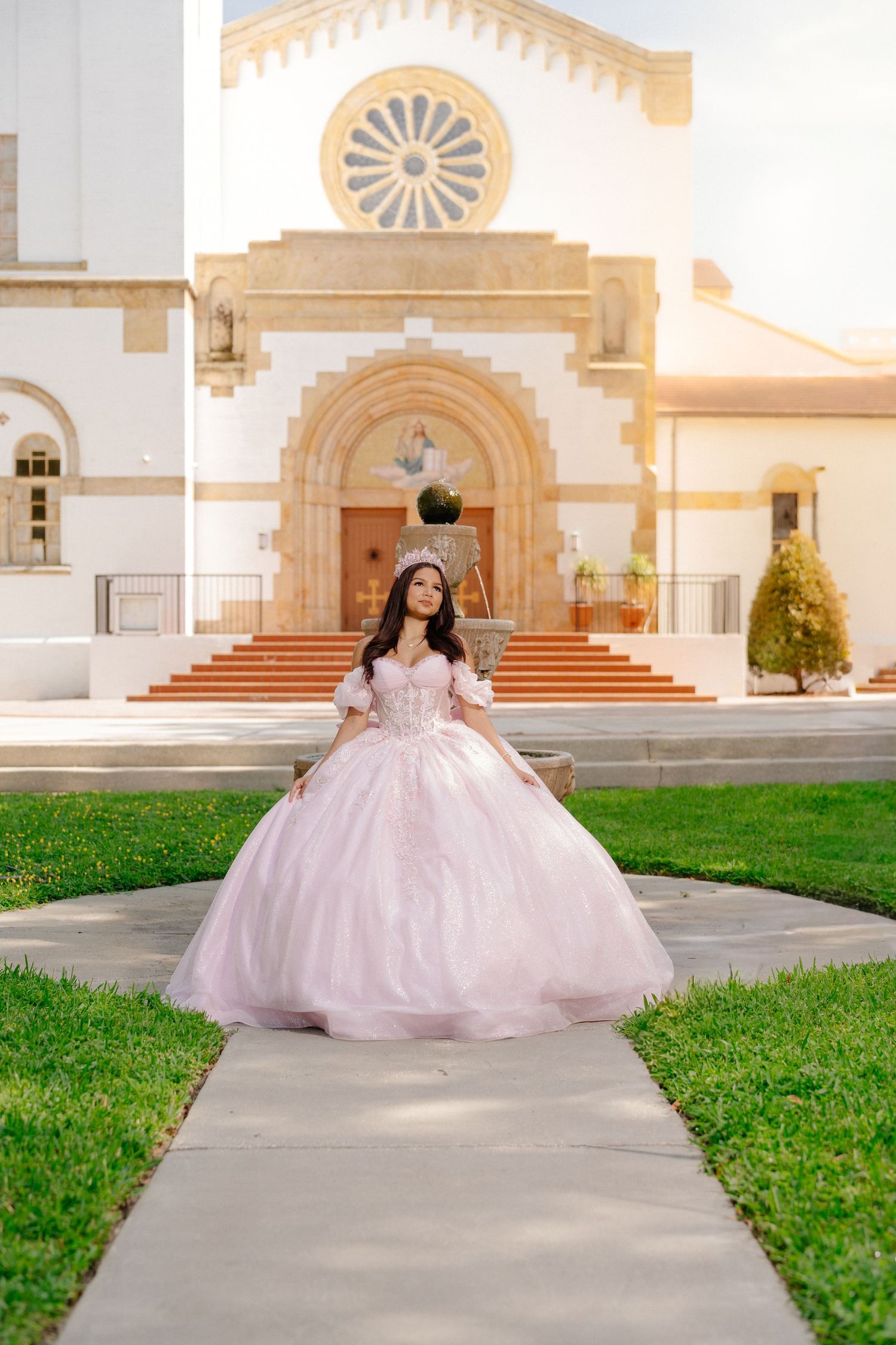 Young woman in pink quinceañera ball gown and tiara poses on church pathway during golden hour.