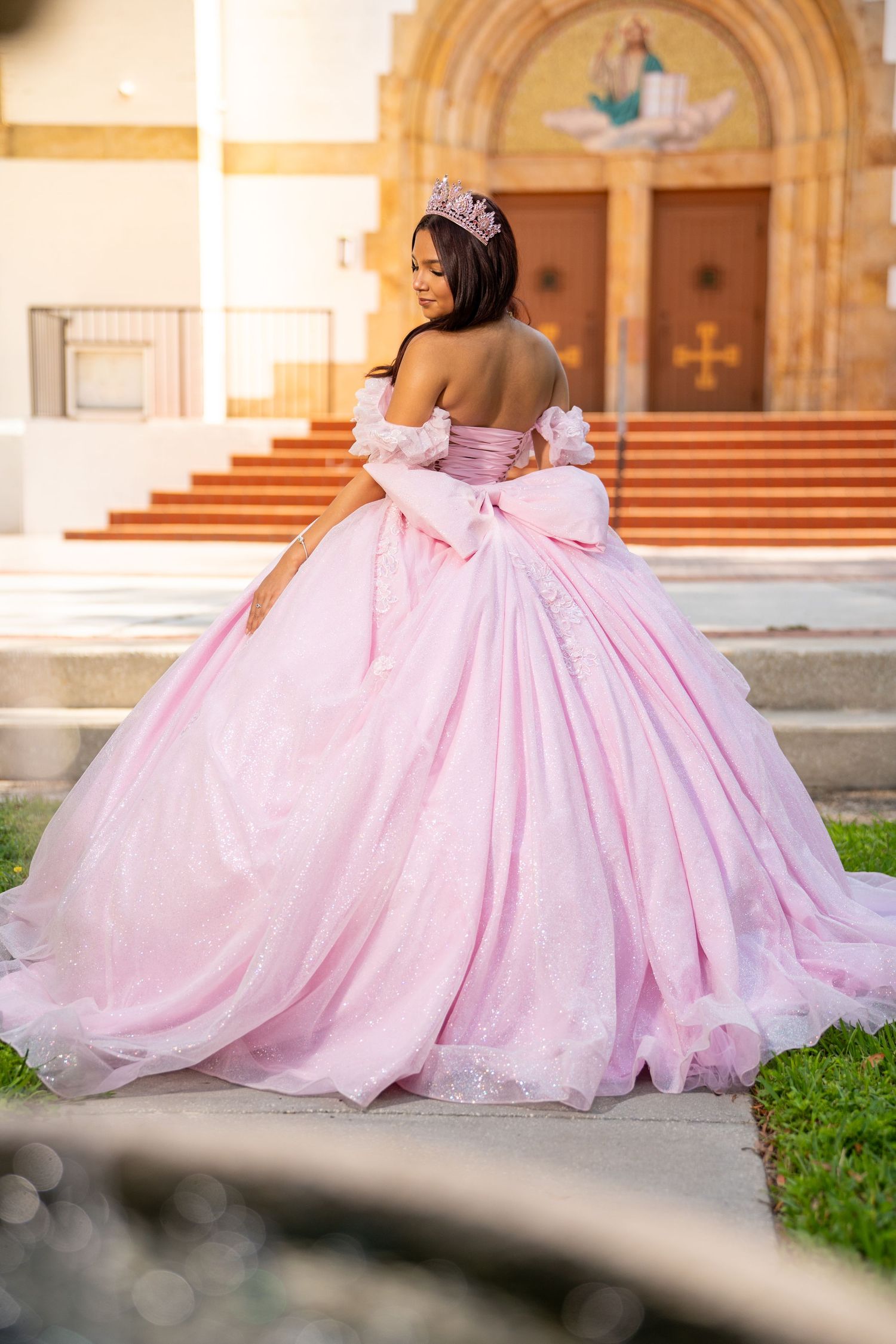 Young woman in a pink ball gown and tiara poses outside a church for her quinceañera celebration.