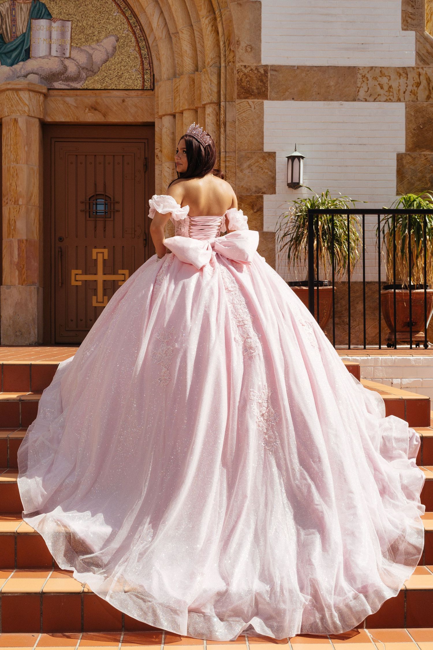 Young woman in pink ball gown with large bow and floral details stands on church steps for quinceañera.