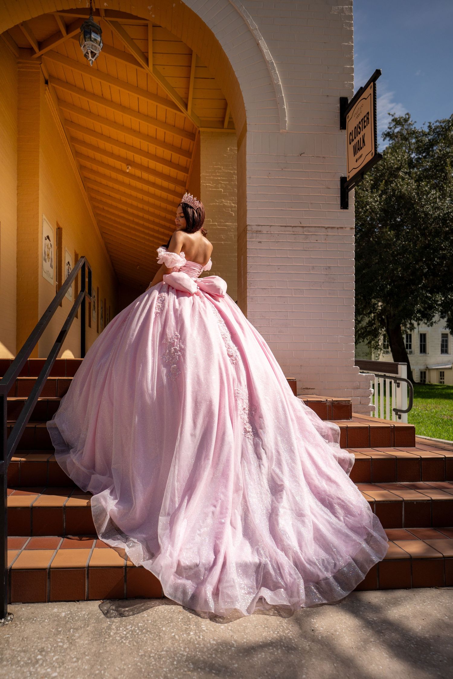 Young woman in a voluminous pink ball gown ascending church steps, viewed from behind, wearing a floral hair piece.