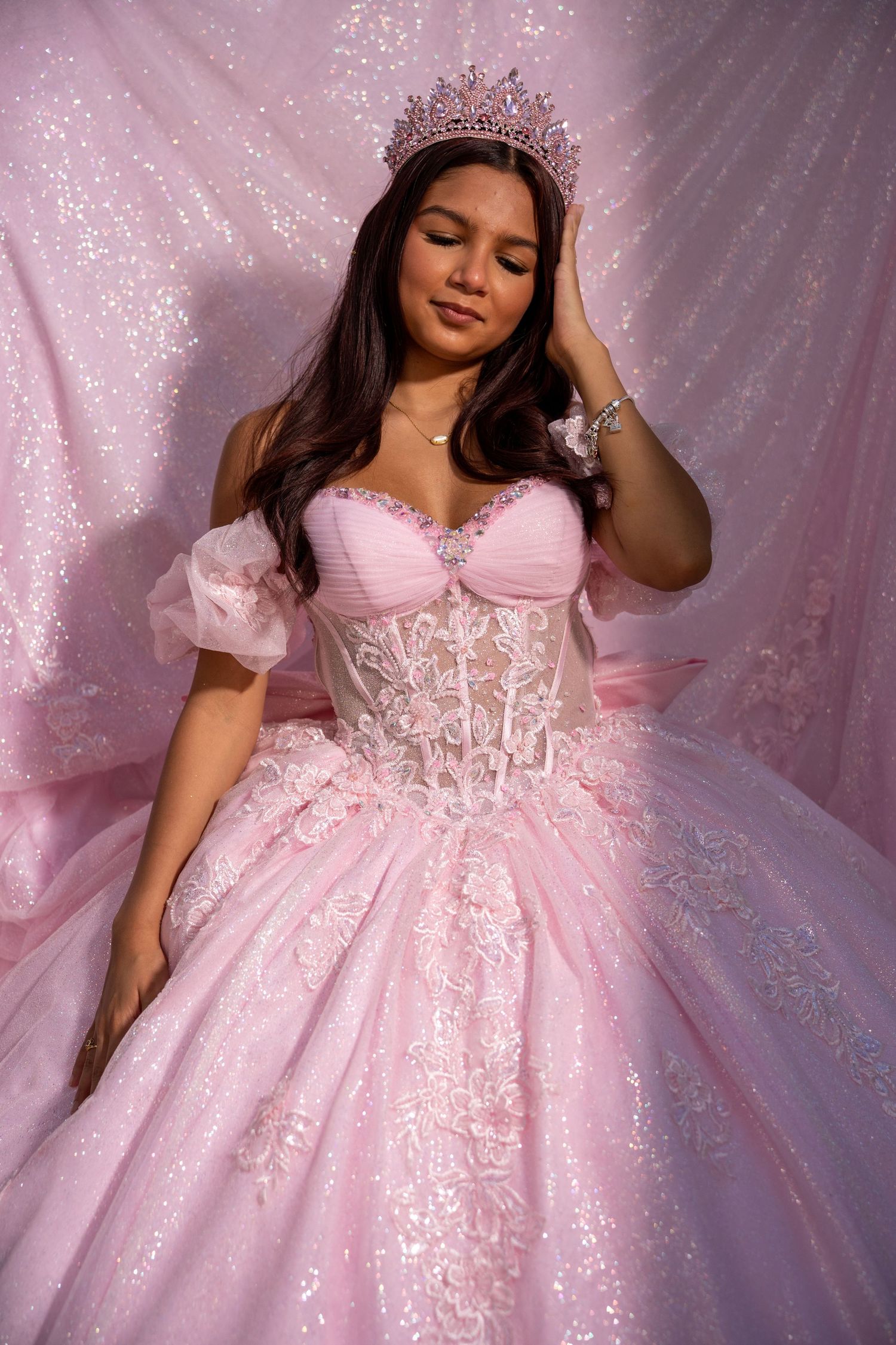 Young woman in pink ball gown with lace corset, puff sleeves, and tiara poses against sparkly pink backdrop.