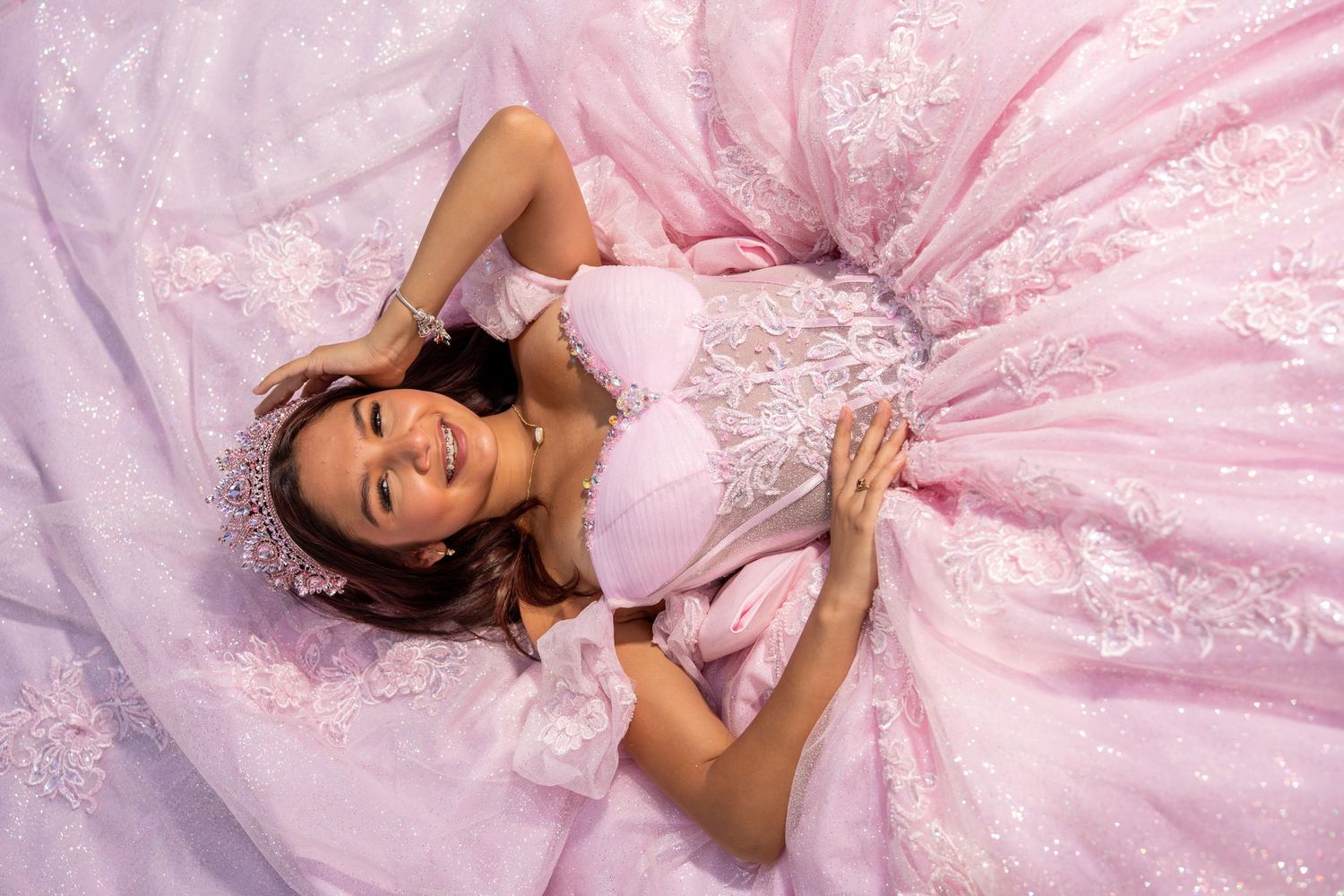 Young woman in elegant pink ball gown with lace details lying down, smiling joyfully at her quinceañera.