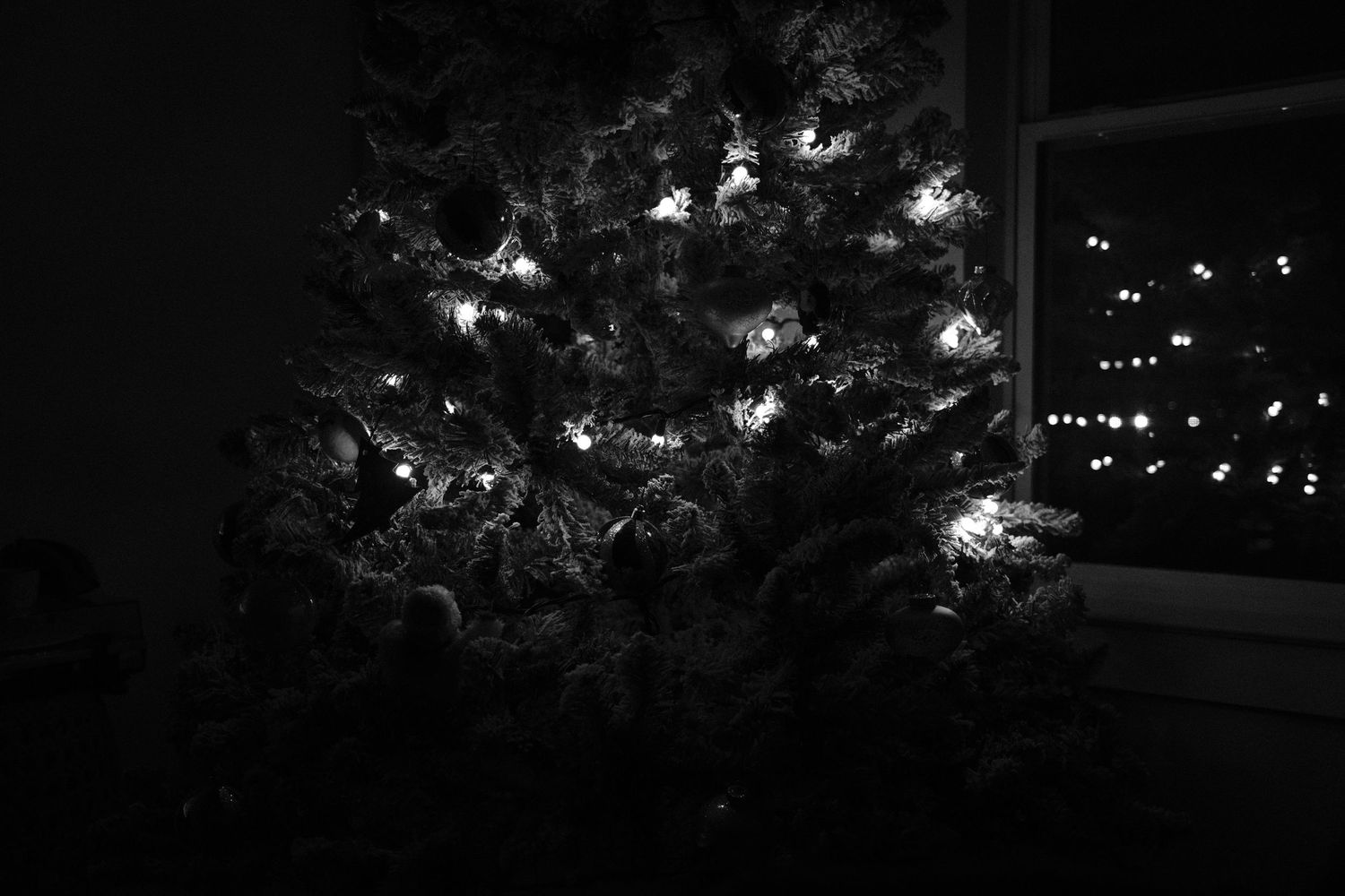 Black and white photo of a decorated Christmas tree glowing with string lights in a dark room near a window.
