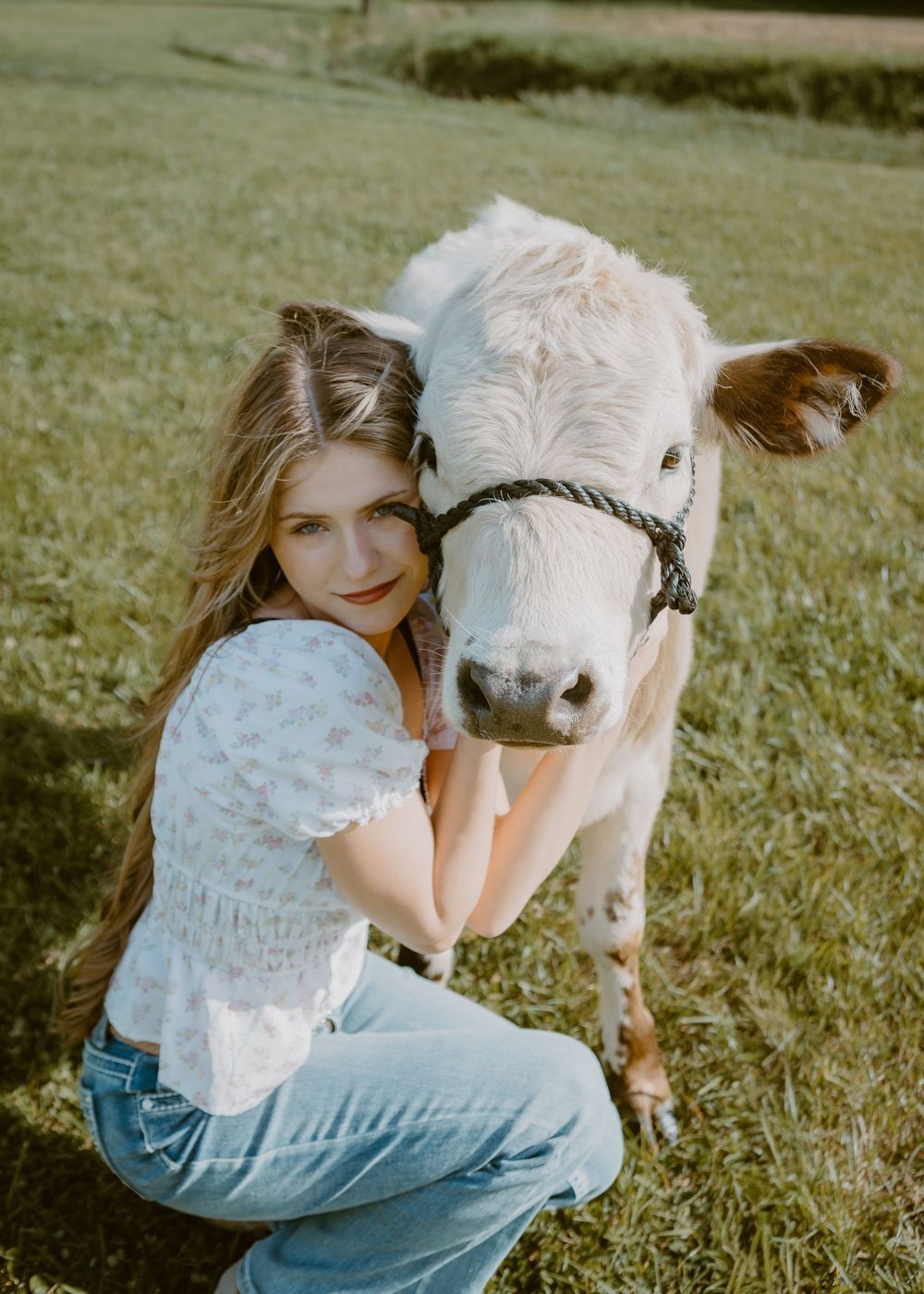 Young woman in white top and jeans crouching beside a white and brown calf on a green grass farm field.