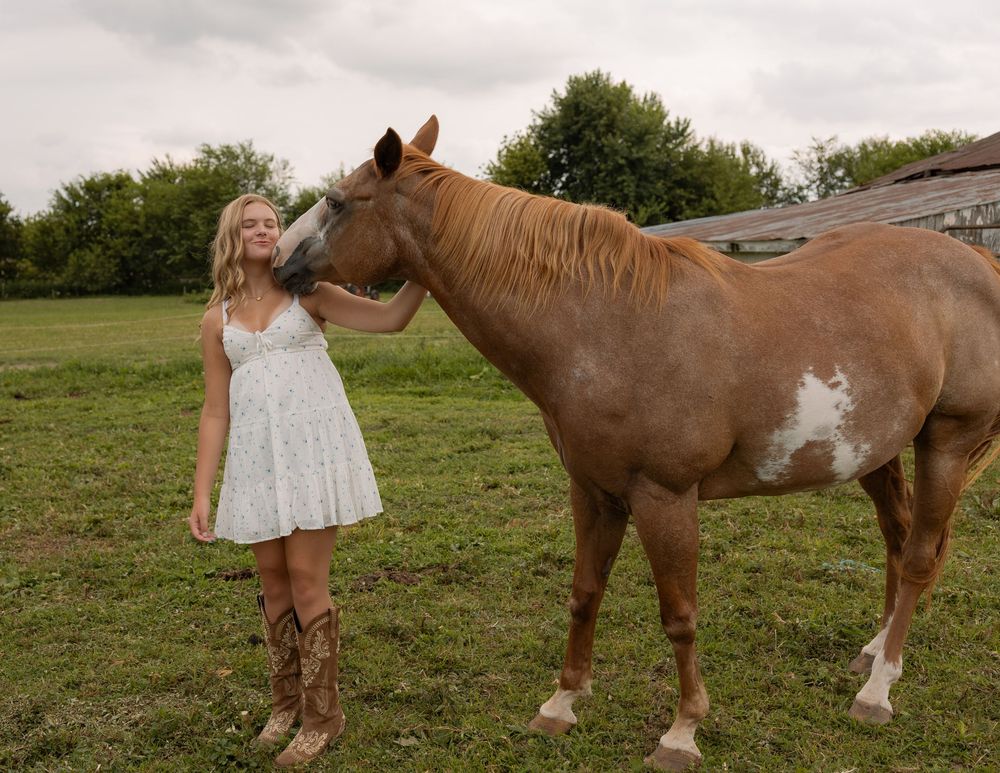 Young blonde woman in white dress and cowboy boots stands beside a brown and white paint horse in a green field.