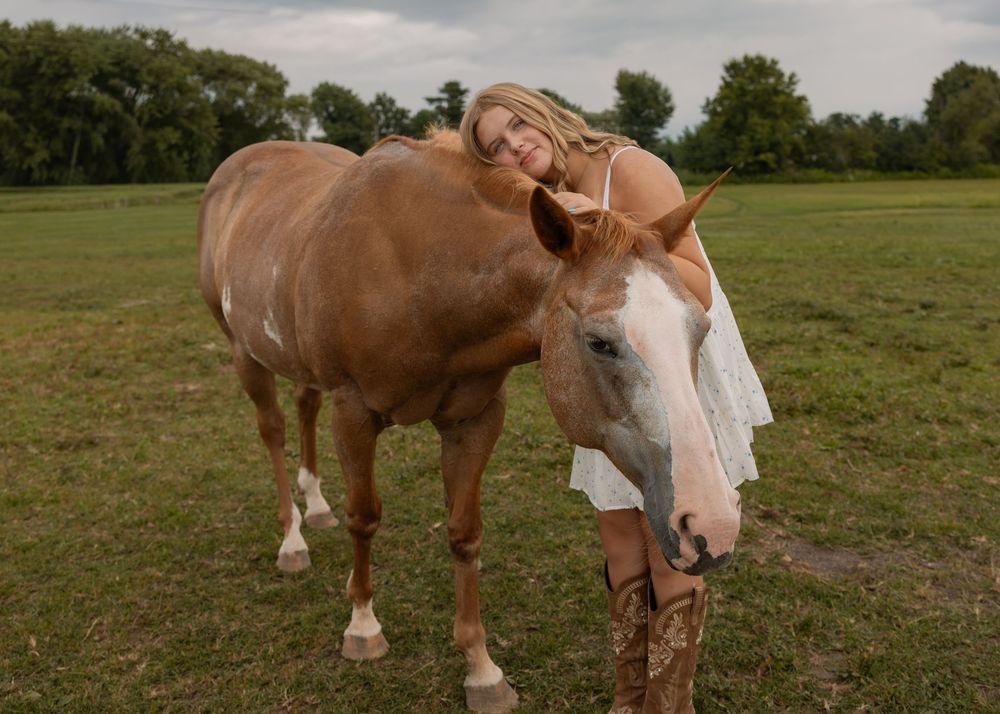 Blonde woman in white dress leaning affectionately on a brown and white horse in a green pasture.