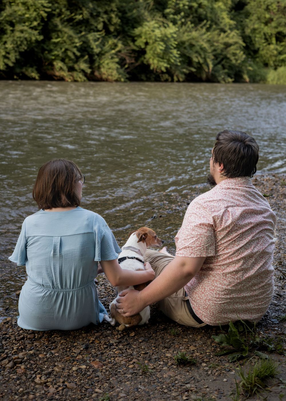 Couple sitting on rocky riverbank with small dog, enjoying peaceful water views surrounded by lush green trees.