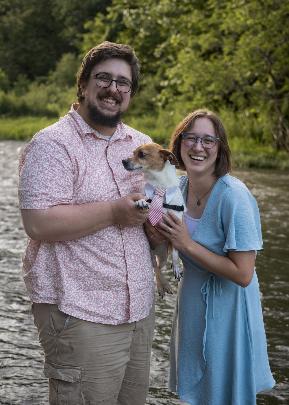 A smiling couple stands by a river holding a small dog wearing a bow tie, surrounded by lush green trees.