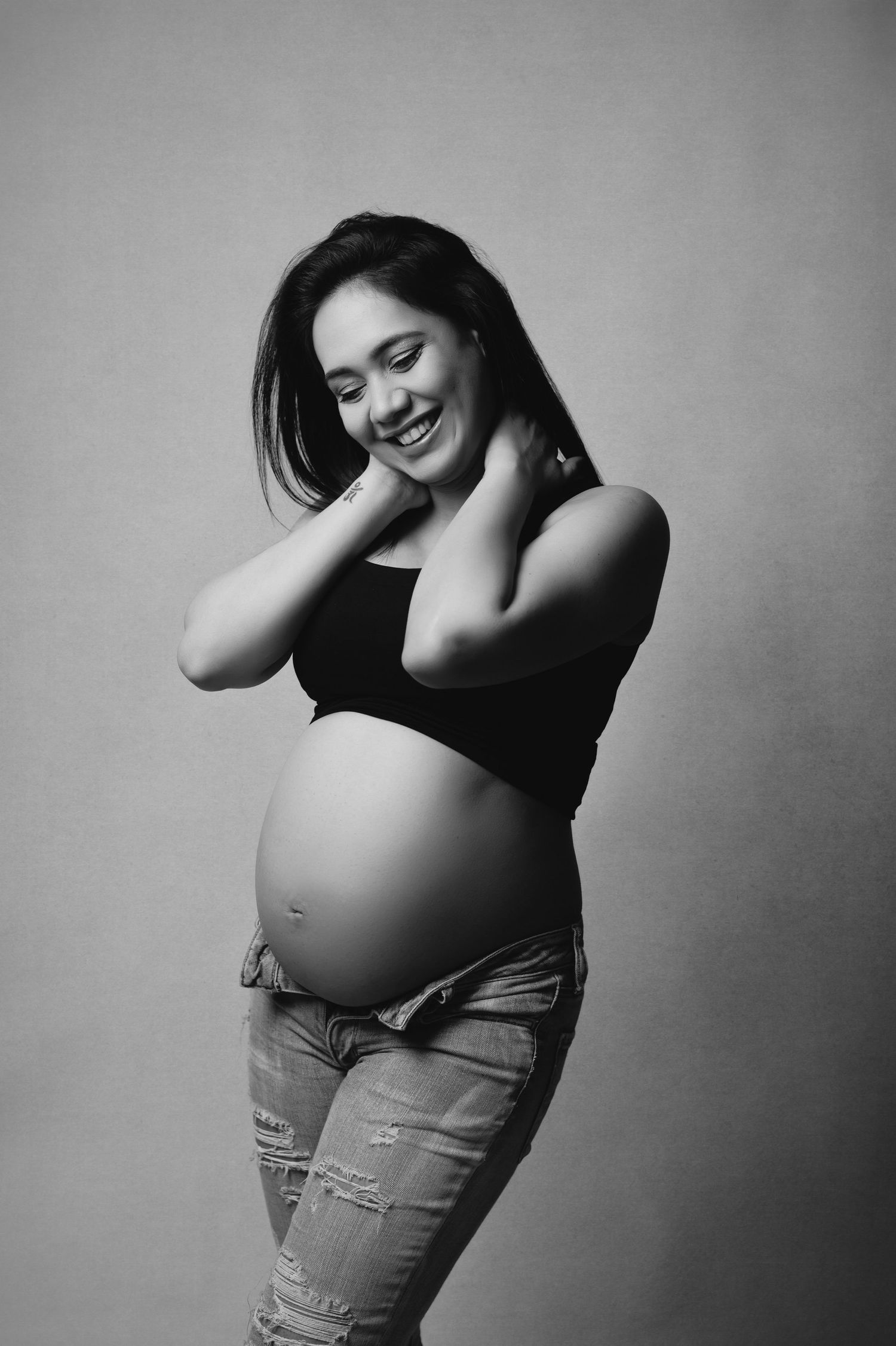 Smiling pregnant woman in black bra and pink robe posing sideways, showing baby bump against dark background.