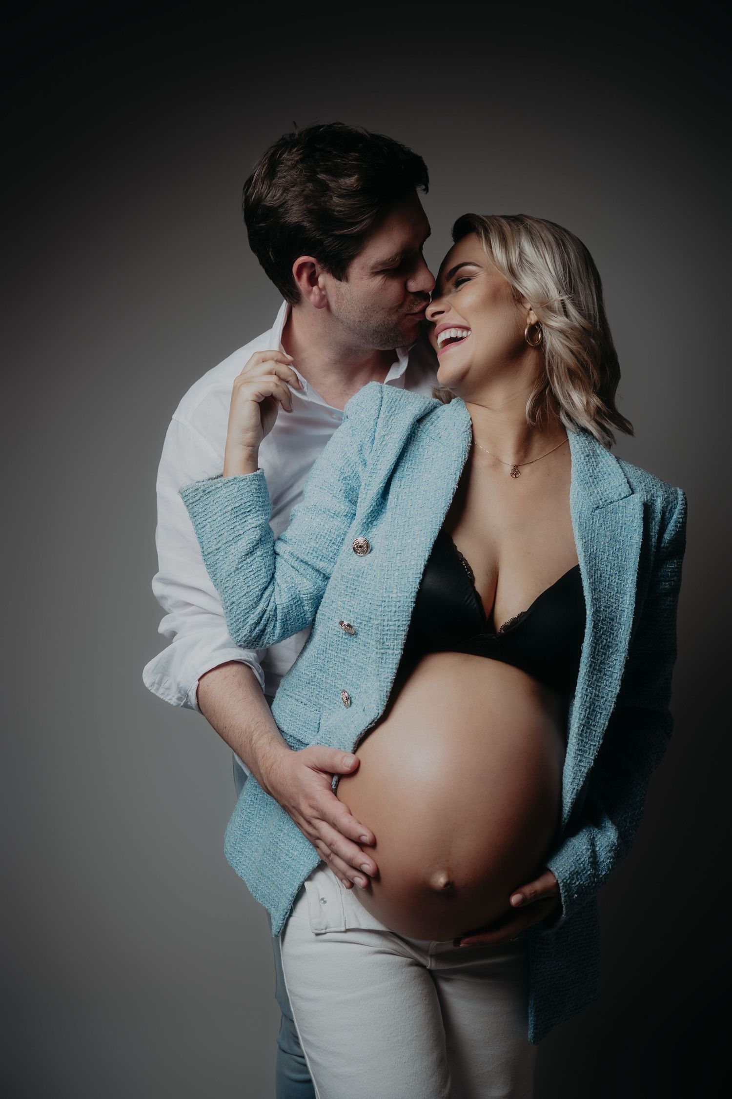 Couple sharing a tender moment, pregnant woman in black bra and blue jacket cradling her baby bump with partner embracing her.