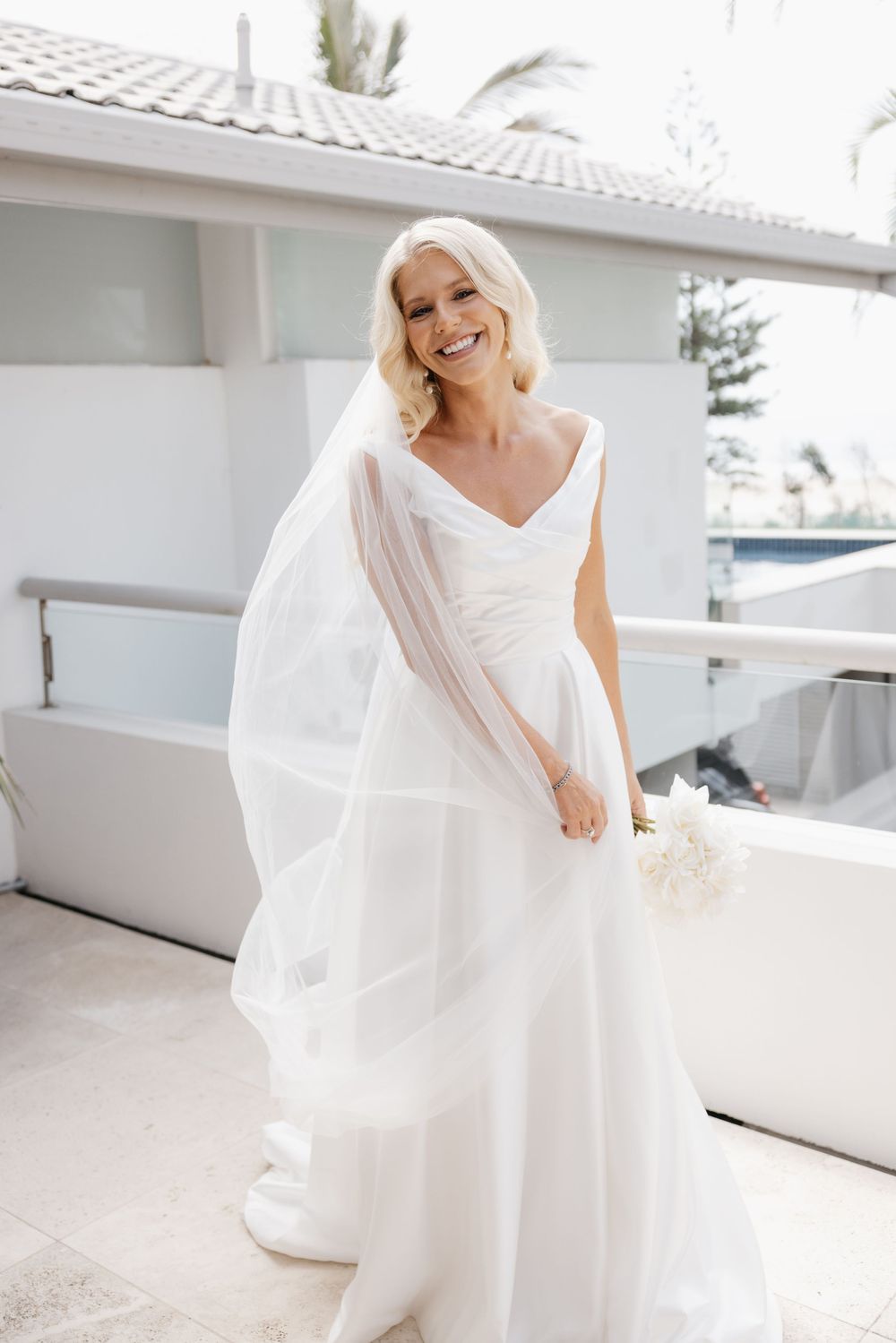 Smiling bride in white A-line gown and veil holding bouquet on sunny outdoor balcony.