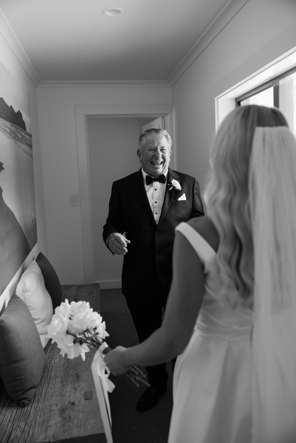 Father in tuxedo laughing joyfully at first look with bride holding white bouquet in elegant wedding dress.