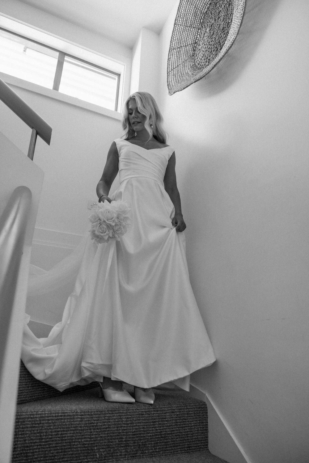 Bride in white gown descending staircase holding bouquet, black and white wedding photo.