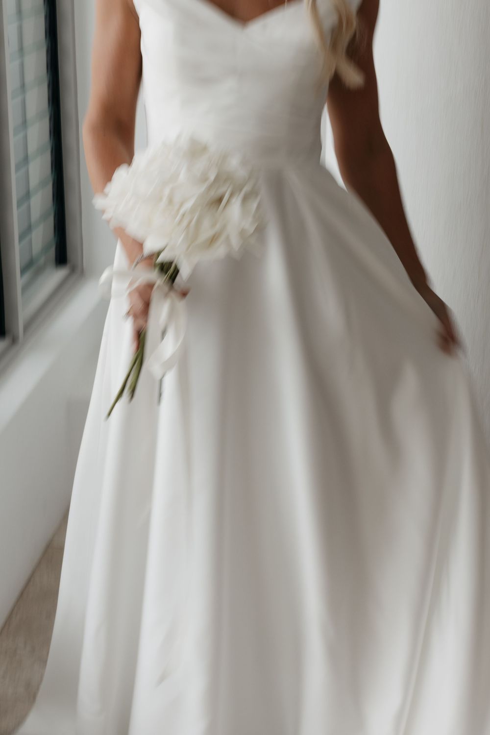 Bride in elegant white satin ball gown holding a white floral bouquet, standing near a window in soft natural light.