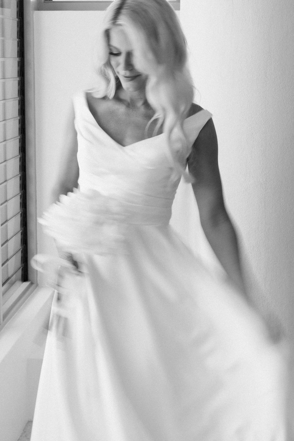 Black and white photo of a smiling blonde bride twirling in an elegant V-neck wedding dress near a window.