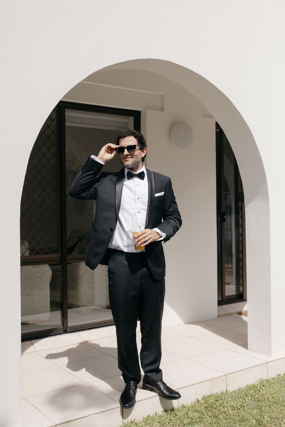 Stylish groom in black tuxedo and bow tie adjusting sunglasses while holding a drink under a white arch.