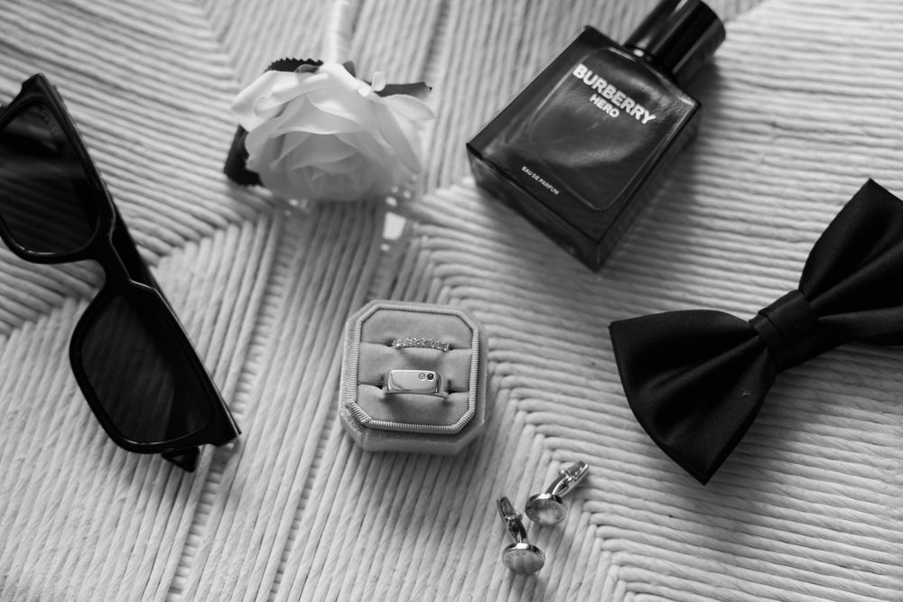 Black and white flat lay of wedding accessories including rings, bow tie, sunglasses, Burberry cologne, and a white flower.