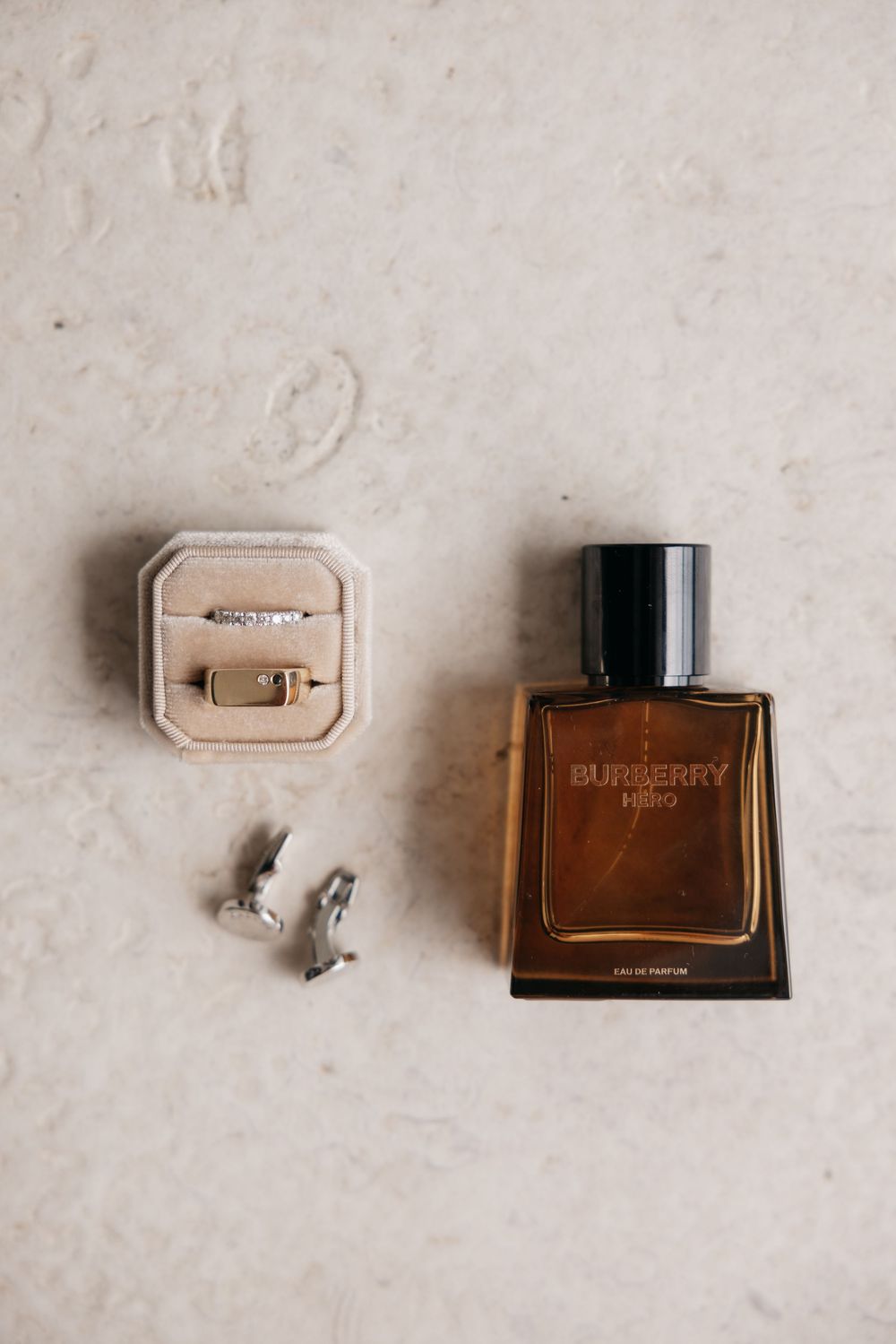 Burberry Hero eau de parfum bottle beside a velvet ring box with wedding bands and airplane cufflinks on marble.