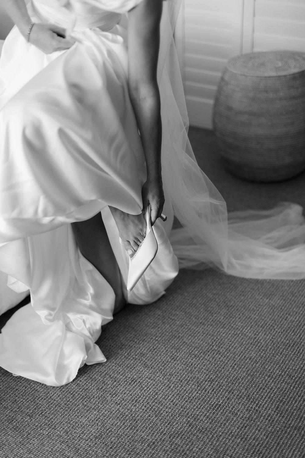 Bride in white satin gown putting on elegant heels, black and white wedding preparation photo.