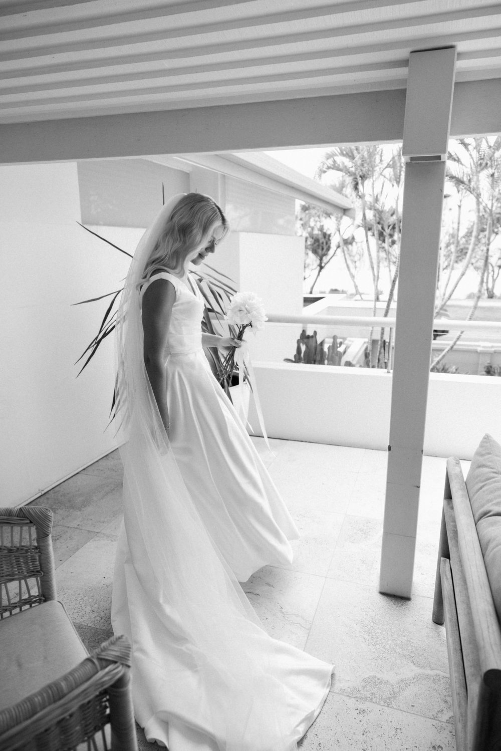 Black and white photo of a bride in a flowing white gown holding a bouquet on a sunlit terrace.