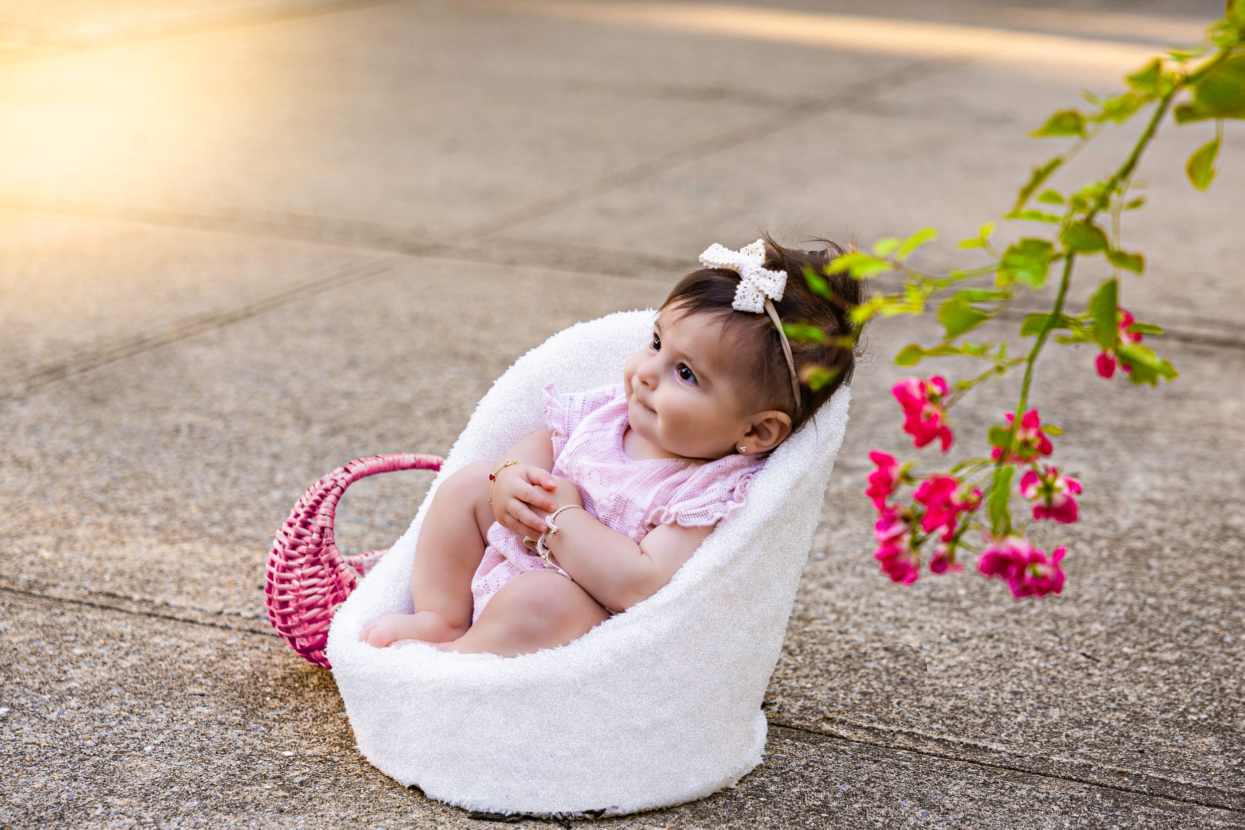 Baby girl in pink outfit sits in white fluffy chair outdoors near red flowers during golden hour.