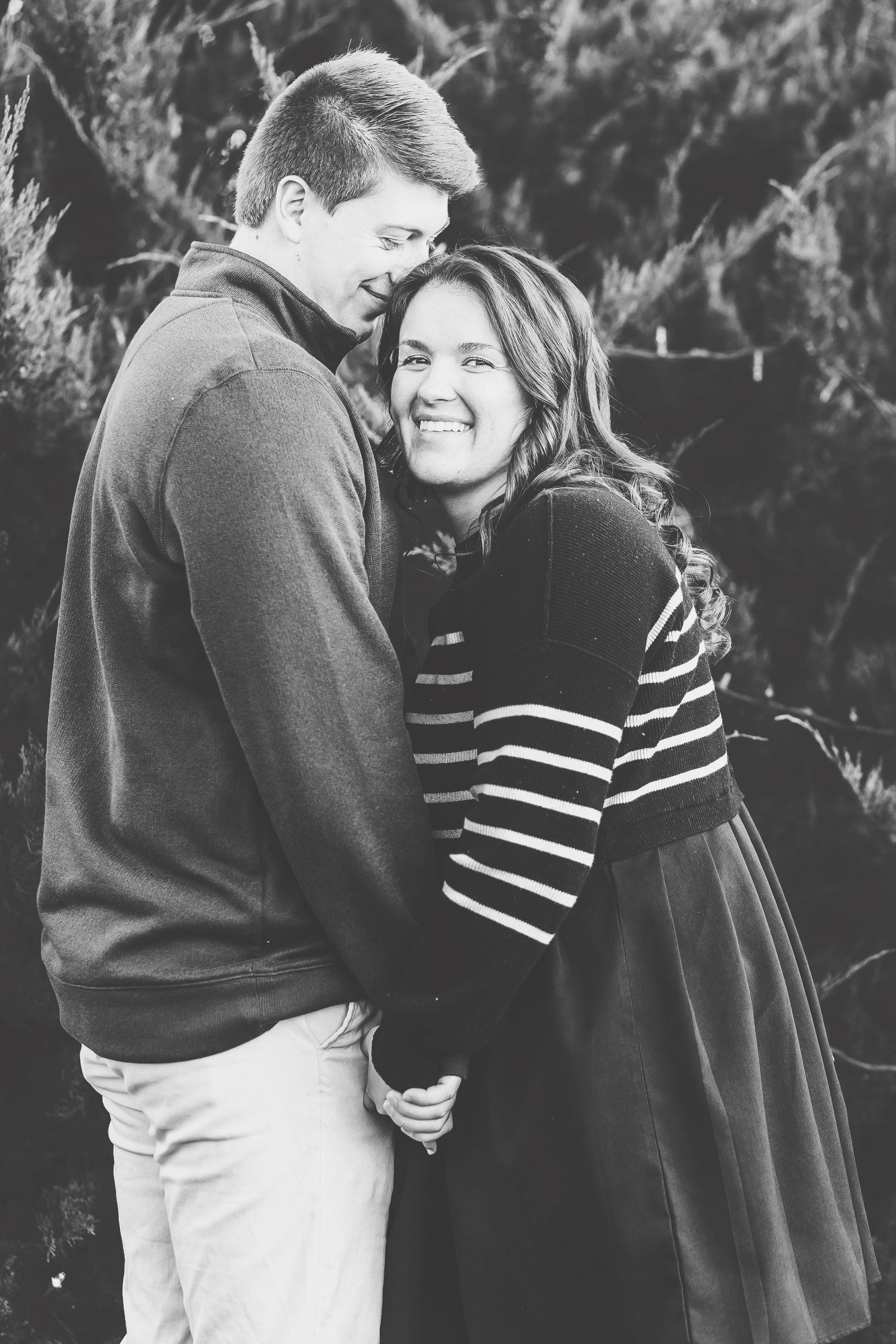 Black and white photo of a smiling couple embracing outdoors, woman in striped dress, man in sweater, among evergreen trees.