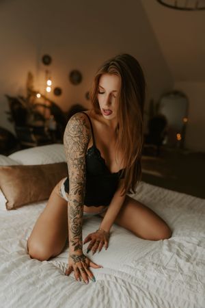 Woman with tattoo sleeve in black lingerie posing on white bed in moody bedroom setting.