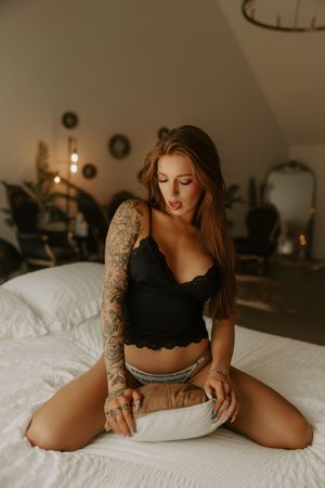 Tattooed woman in black lace crop top and denim shorts sitting on white bed in boho bedroom.