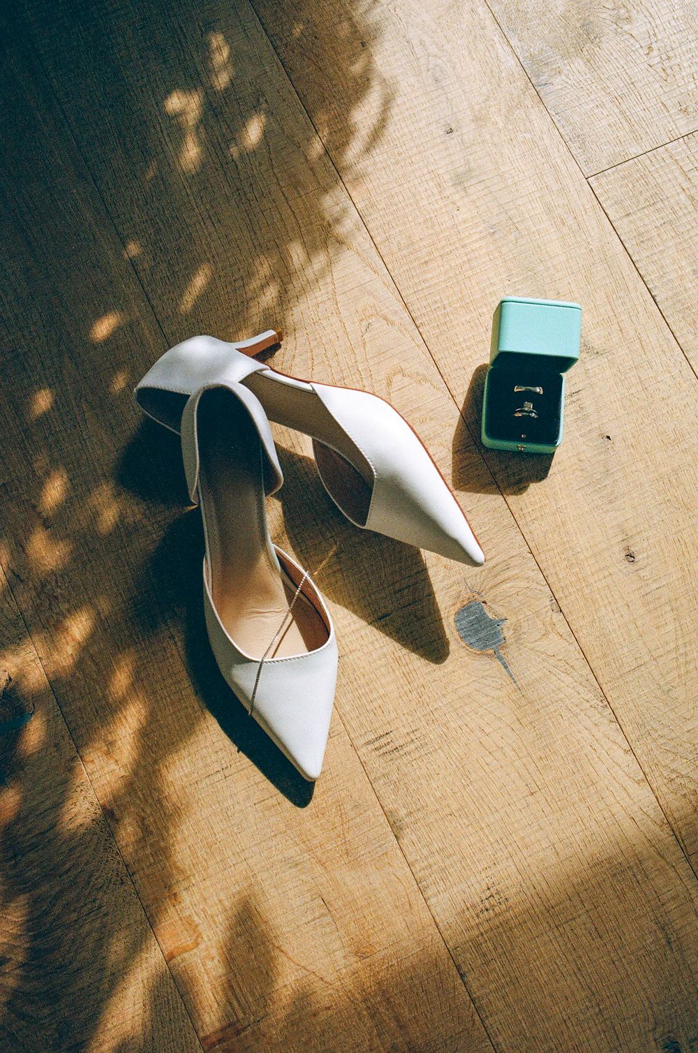 White bridal heels and a mint green ring box on a wooden floor with dappled sunlight.
