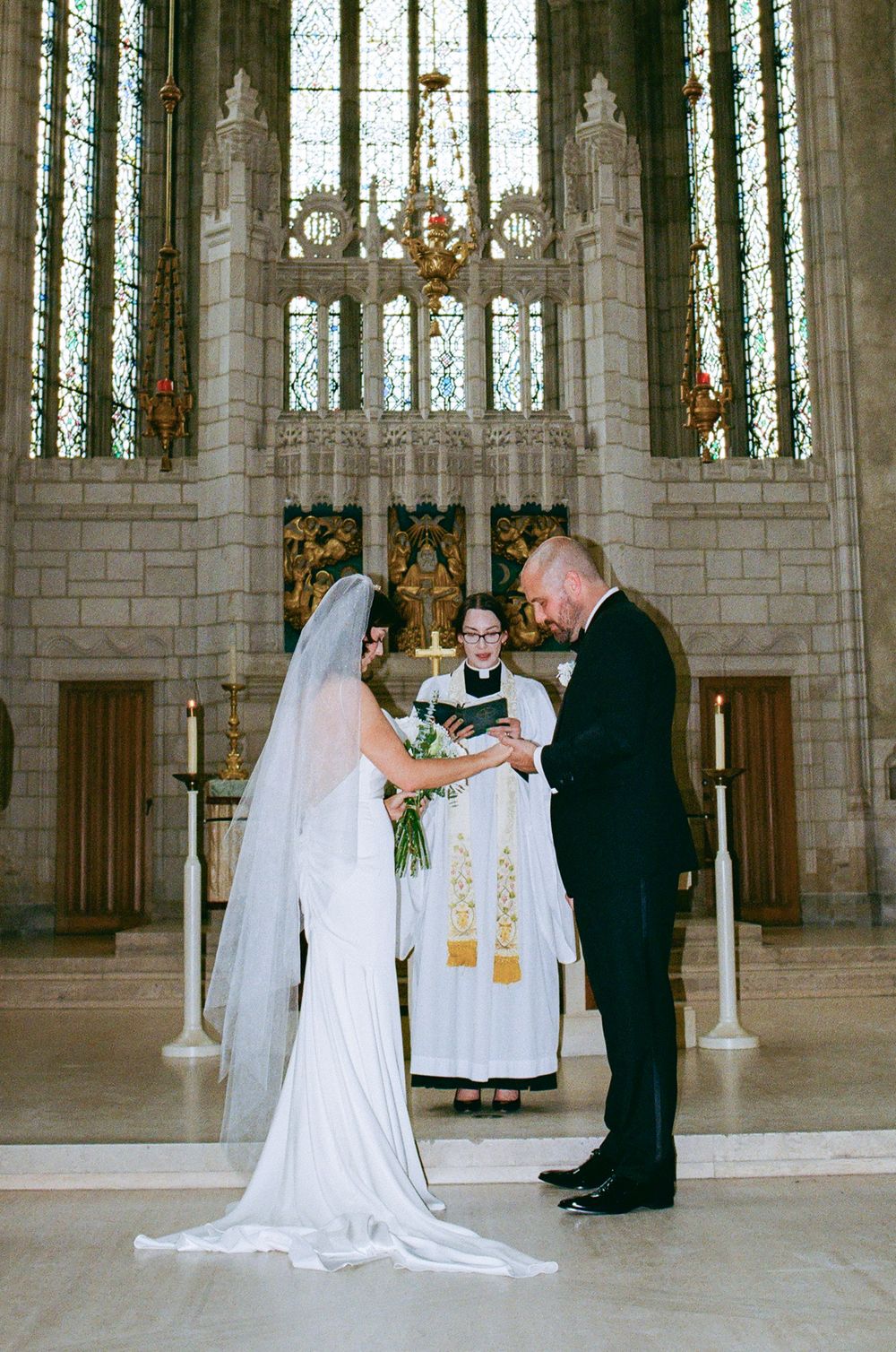 Bride and groom exchange vows at ornate church altar with stained glass windows and officiant in white robes.
