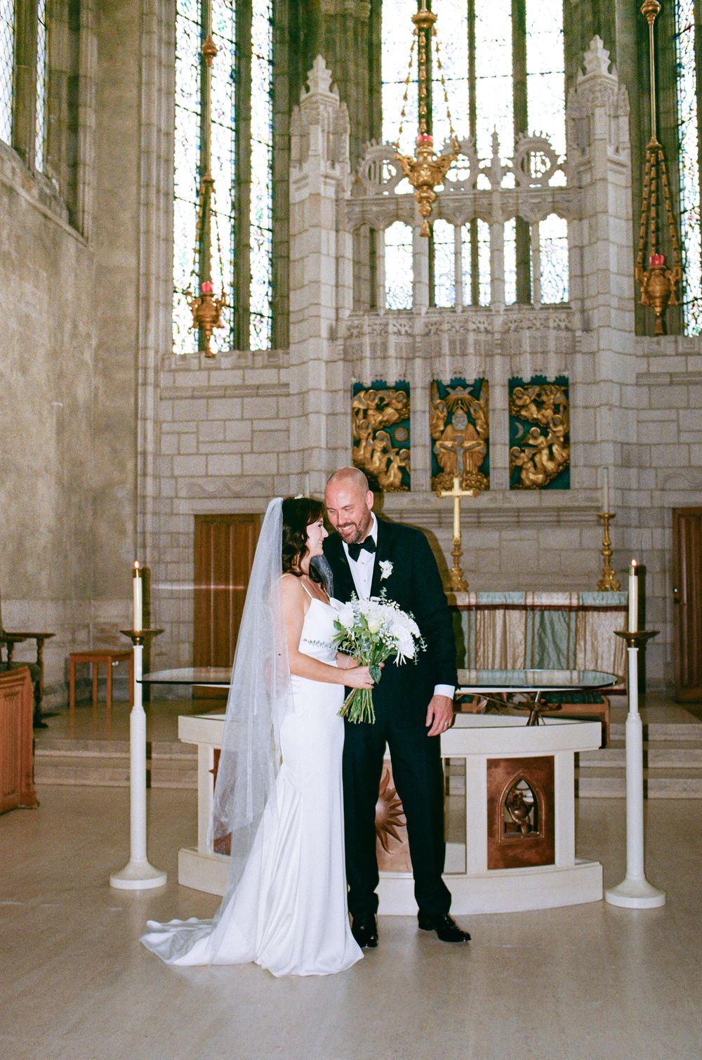 Bride and groom share a romantic moment at a church altar, bride in white gown with veil holding a bouquet.