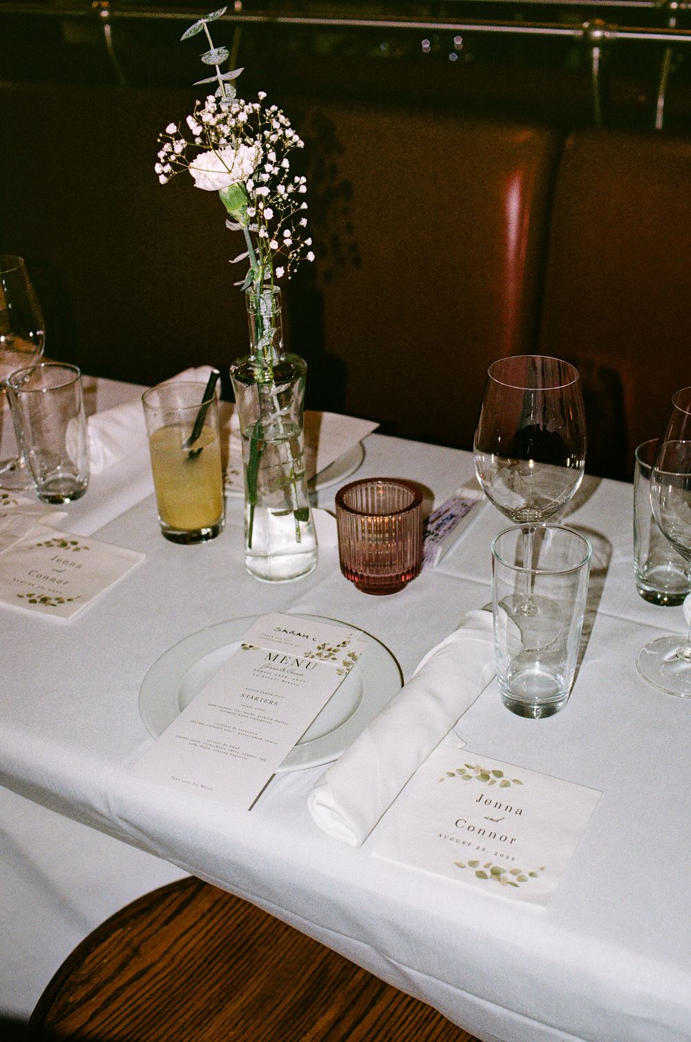 Elegant wedding reception table setting with floral centerpiece, menu cards, wine glasses, and candles on white linen.