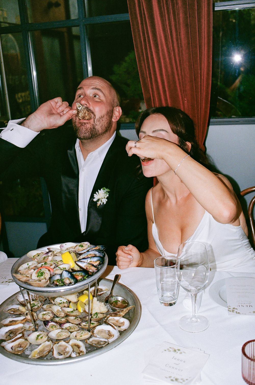 Bride and groom joyfully eating oysters at their wedding reception dinner table with a seafood tower.