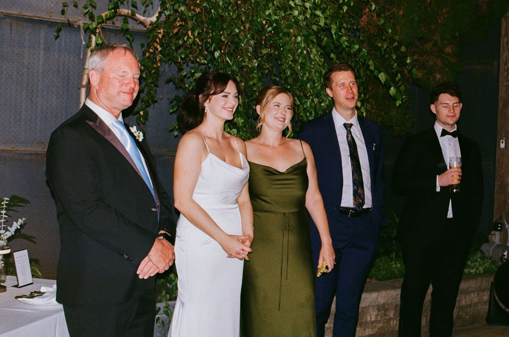 Wedding guests in formal attire smile together indoors, including a bride in white and guests in navy and olive green.