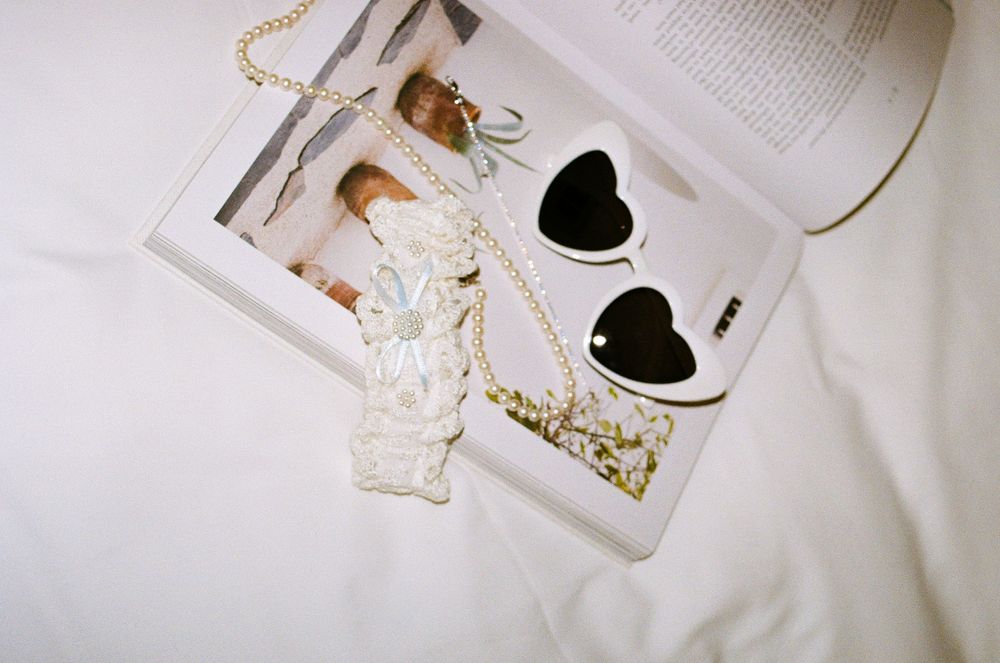 Heart-shaped black sunglasses and pearl chain necklace resting on an open fashion magazine on white bedding.