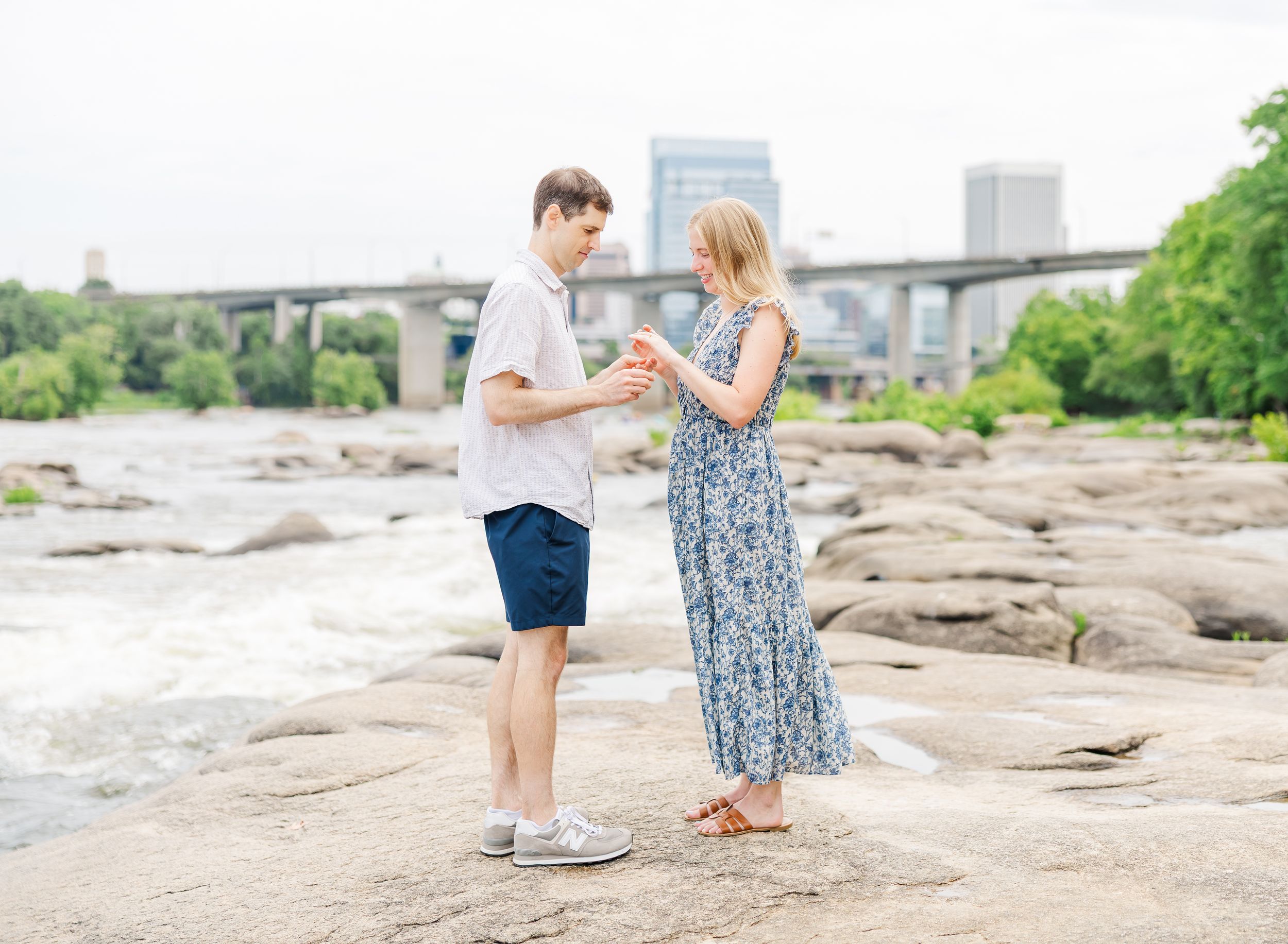 Couple holding hands on rocky riverbank with city bridge and skyline in background on cloudy day.