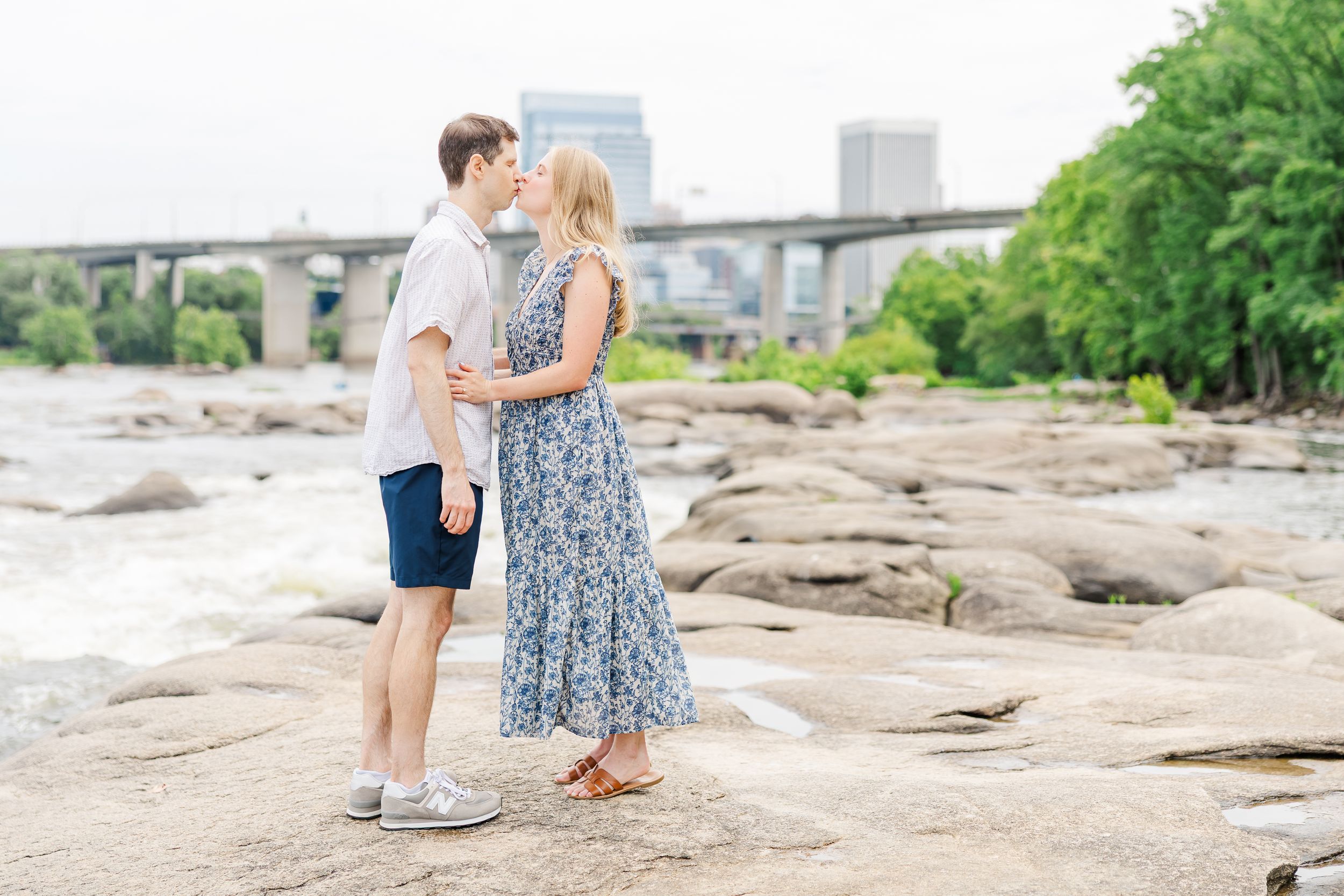 Couple sharing a romantic kiss on rocky riverbank with bridge and city skyline in background.
