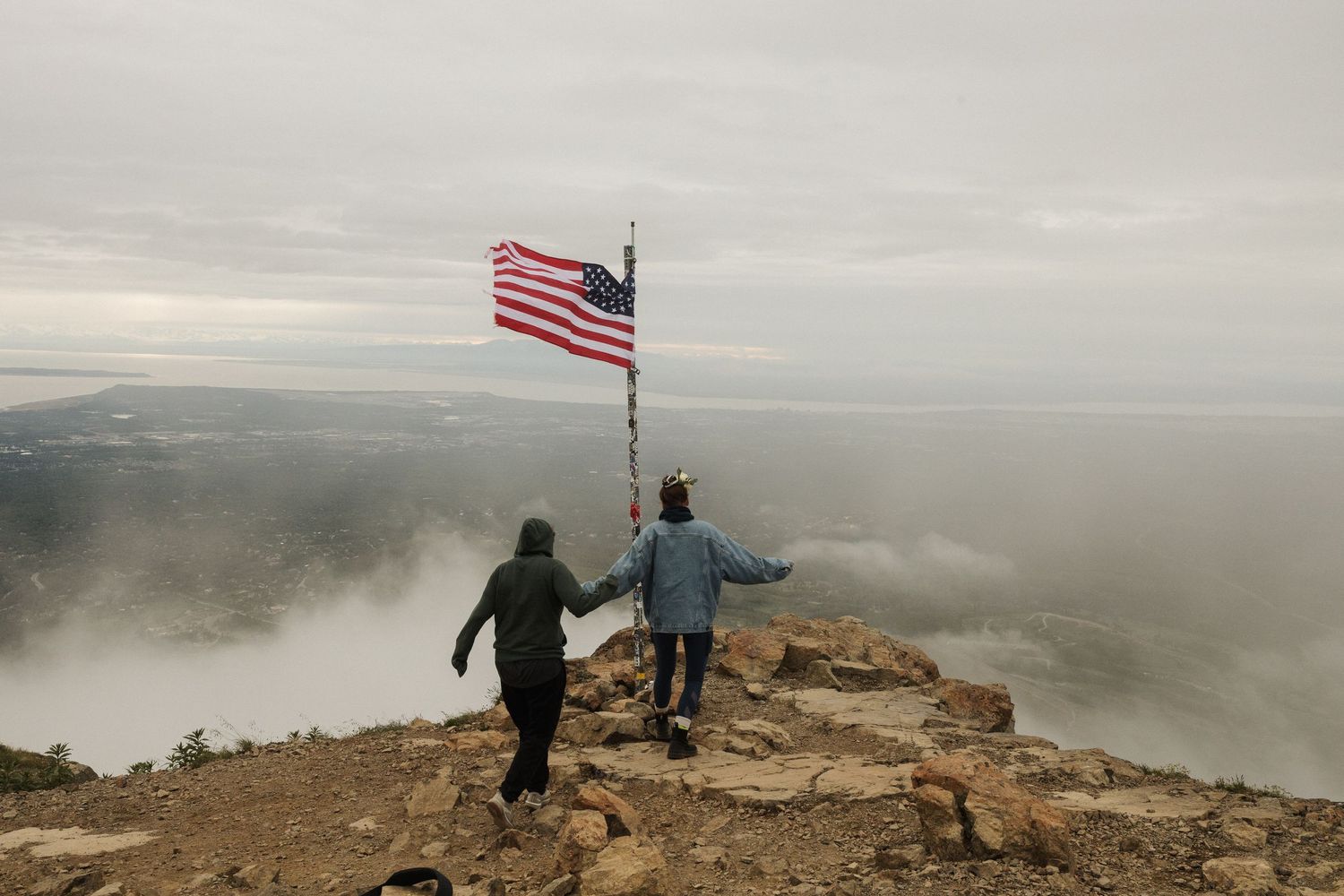 Two people holding hands near an American flag on a misty mountain summit with scenic views in Alaska.