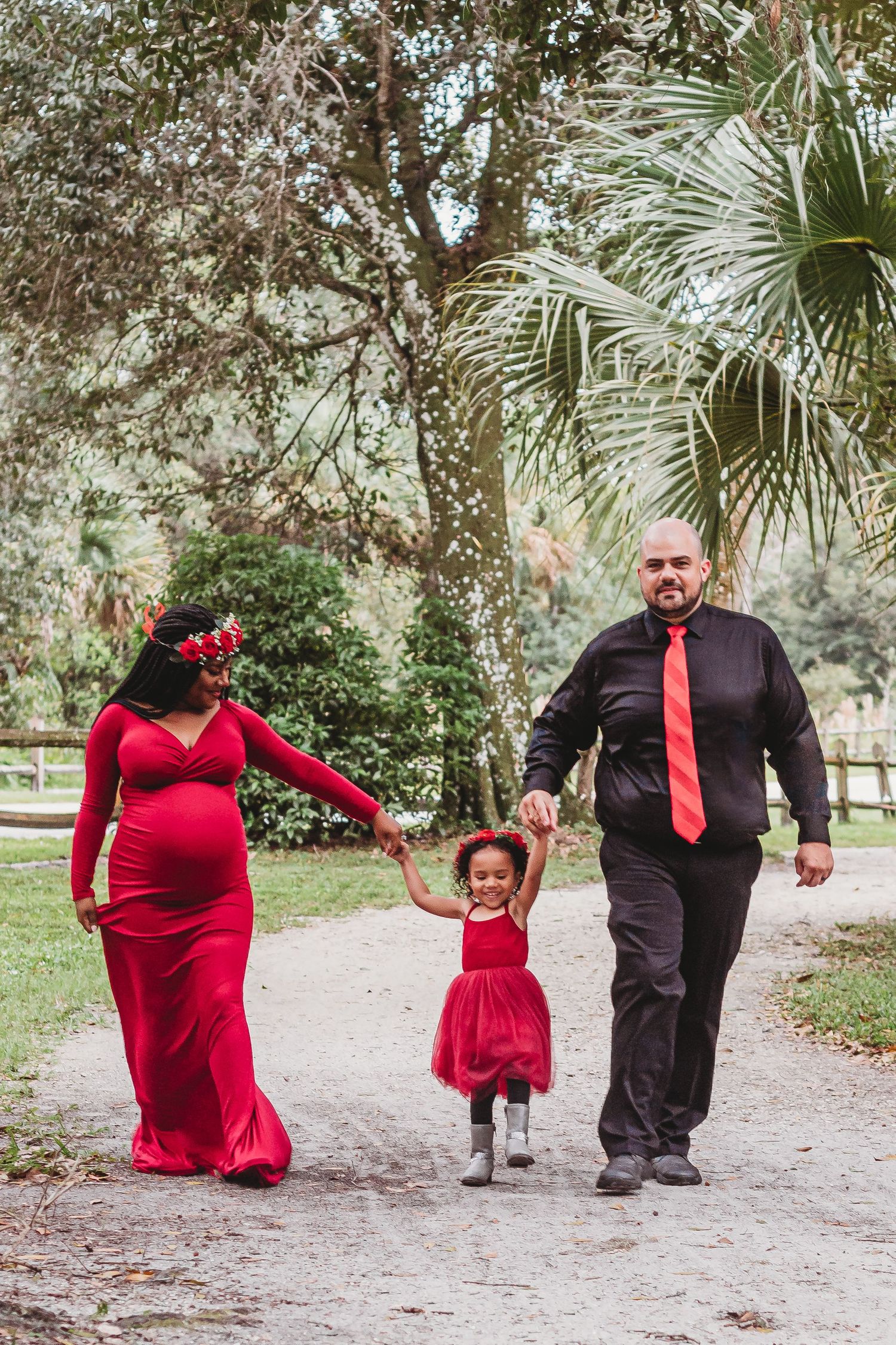 Pregnant woman in red gown walks with husband in black suit and young daughter in red dress along a park path at riverbend park Jupiter.